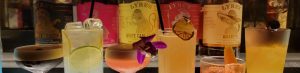 A selection of colorful cocktails in various glasses, garnished with fruit and flowers, with spirit bottles in the background.