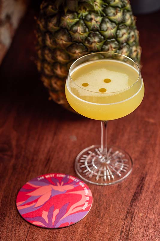 A coupe glass with a yellow cocktail featuring three drops on top sits on a wooden surface next to a colorful coaster and a whole pineapple.