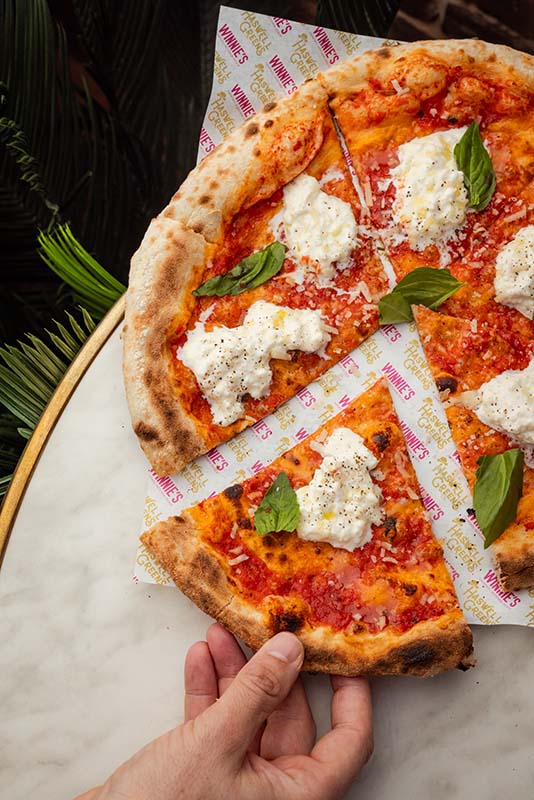 A hand takes a slice of pizza topped with tomato sauce, ricotta cheese, basil leaves, and pepper, served on branded paper on a marble table.