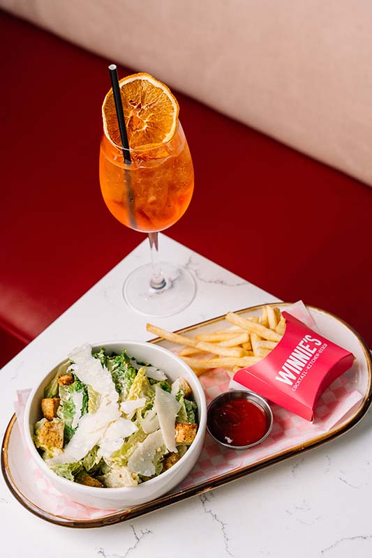 A bowl of Caesar salad, a serving of fries with ketchup, and an orange cocktail garnished with a dried orange slice on a white table.