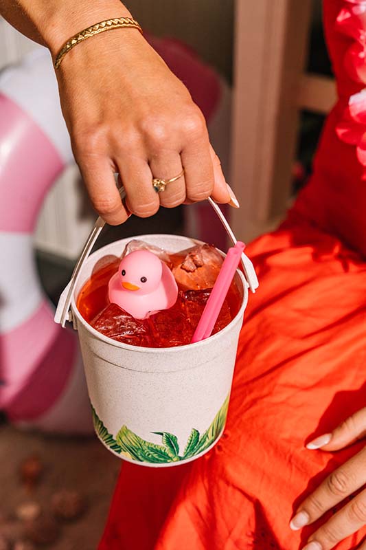 A hand holds a small white bucket filled with a red drink, ice, a pink straw, and a floating pink rubber duck. The person is wearing a red dress.
